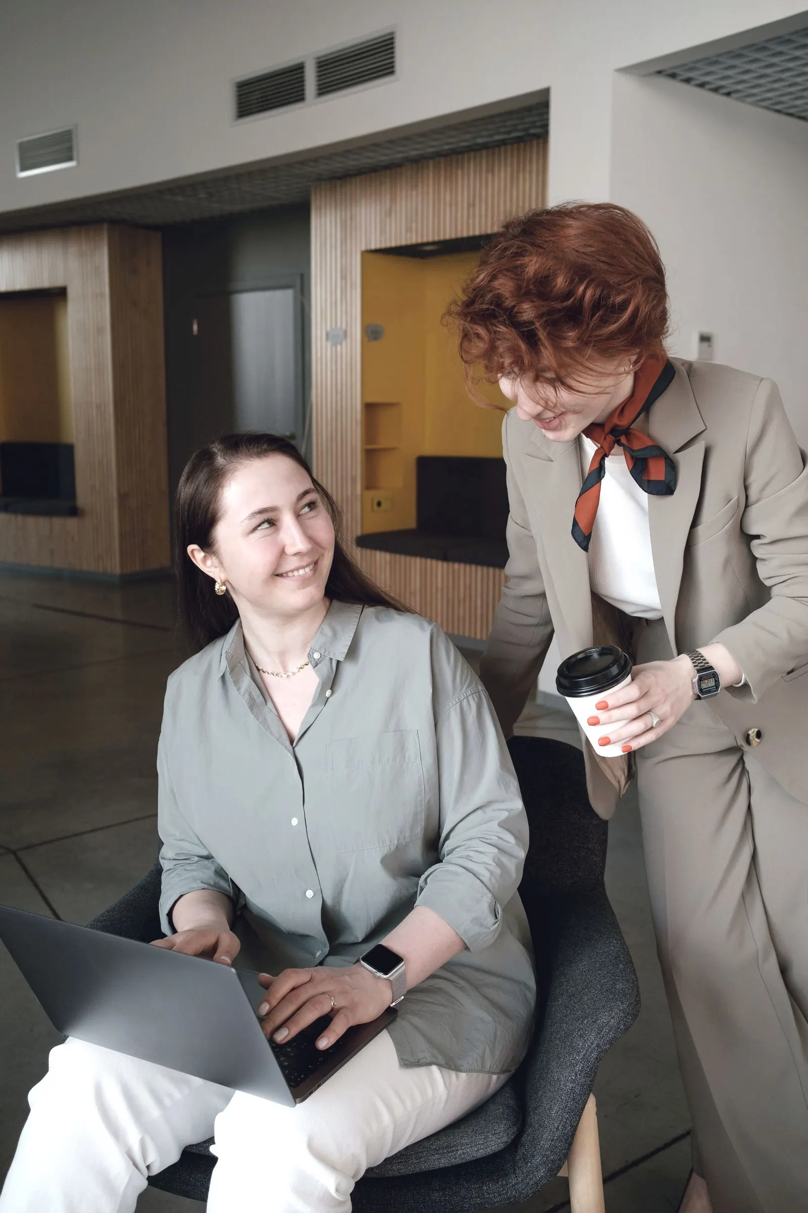 A colleague stands beside a seated coworker with a laptop, talking.