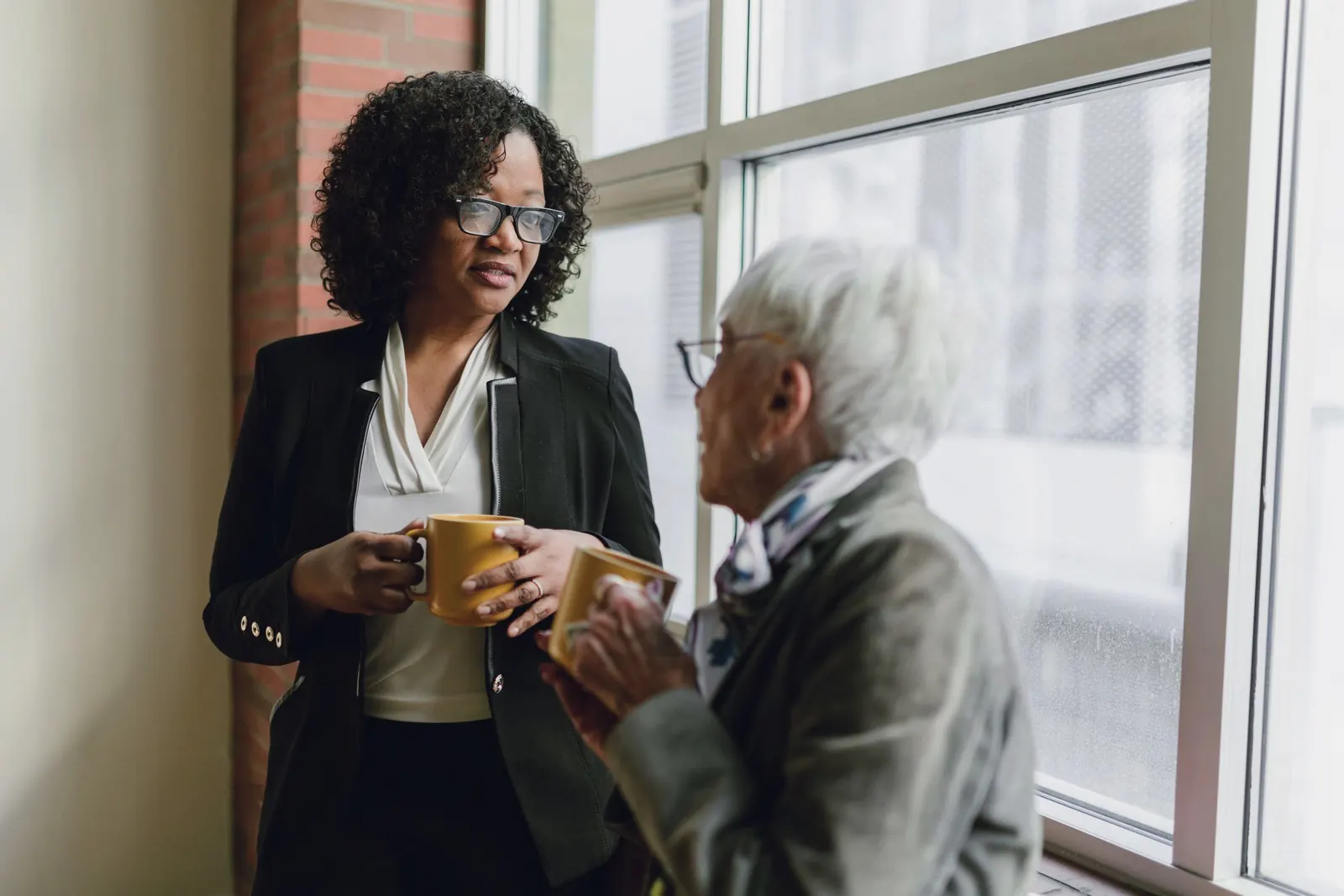 Two colleagues, one older and one younger, talk over mugs beside a bright window.