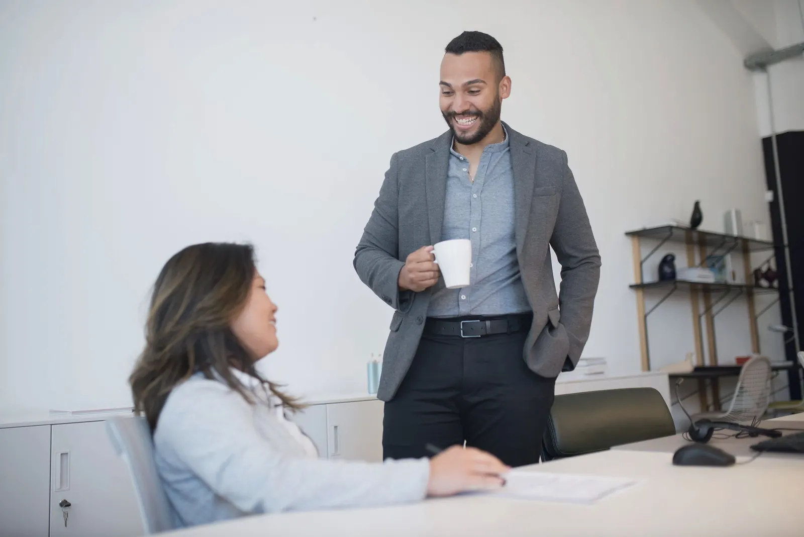 A manager stands with a mug and talks with a colleague seated at a white desk.