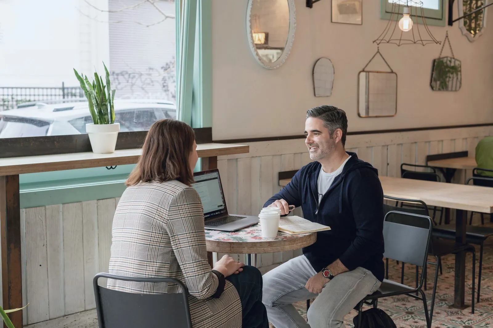 Two colleagues talk a plan through at a cafe table by a window.