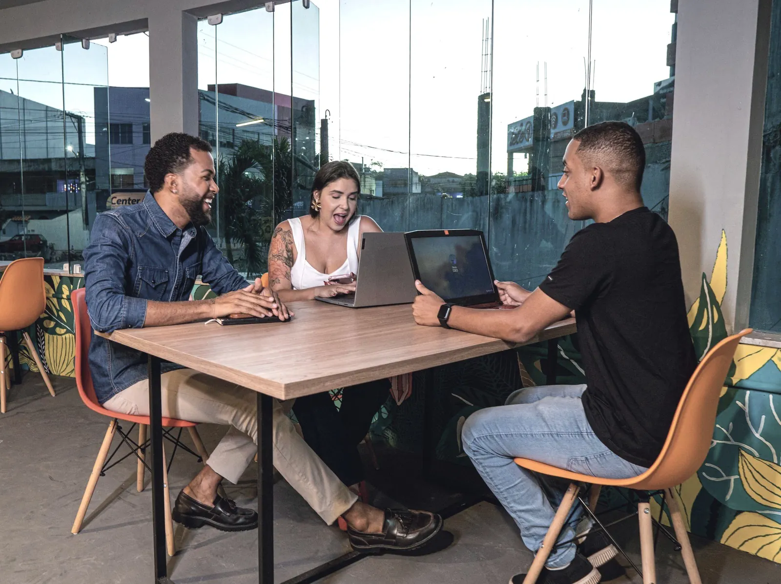 Three colleagues smile as they work on laptops at a table with a view.