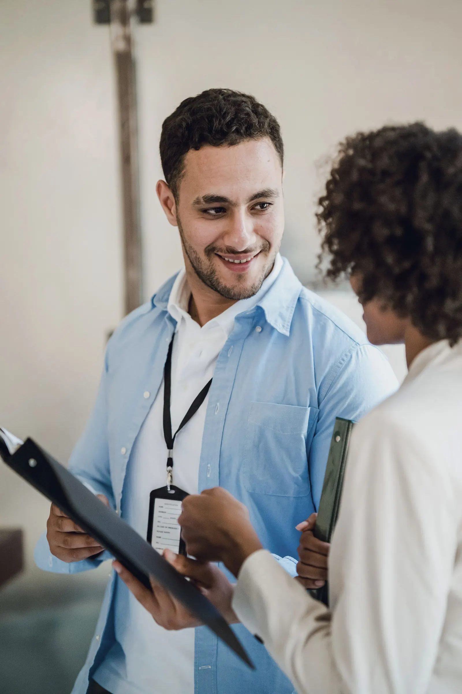 A colleague with a lanyard and a clipboard smiles as he talks with a teammate in a bright office.
