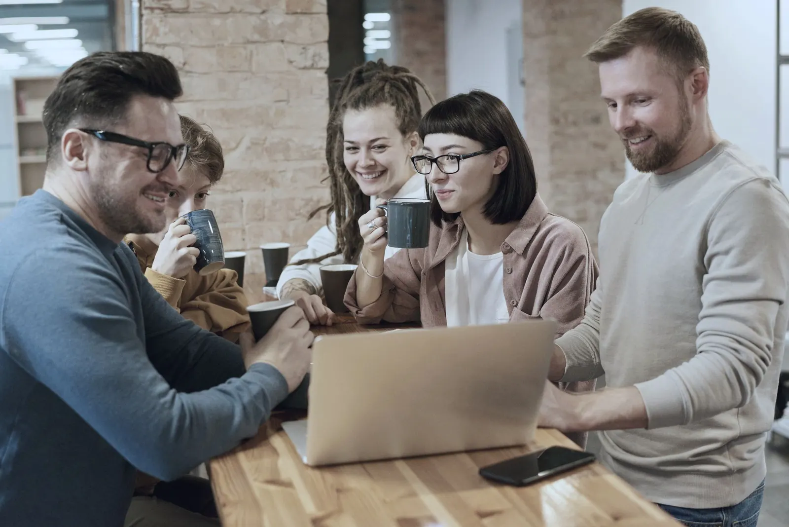 A group shares coffee and a laptop at a wooden table in a brick-walled office.