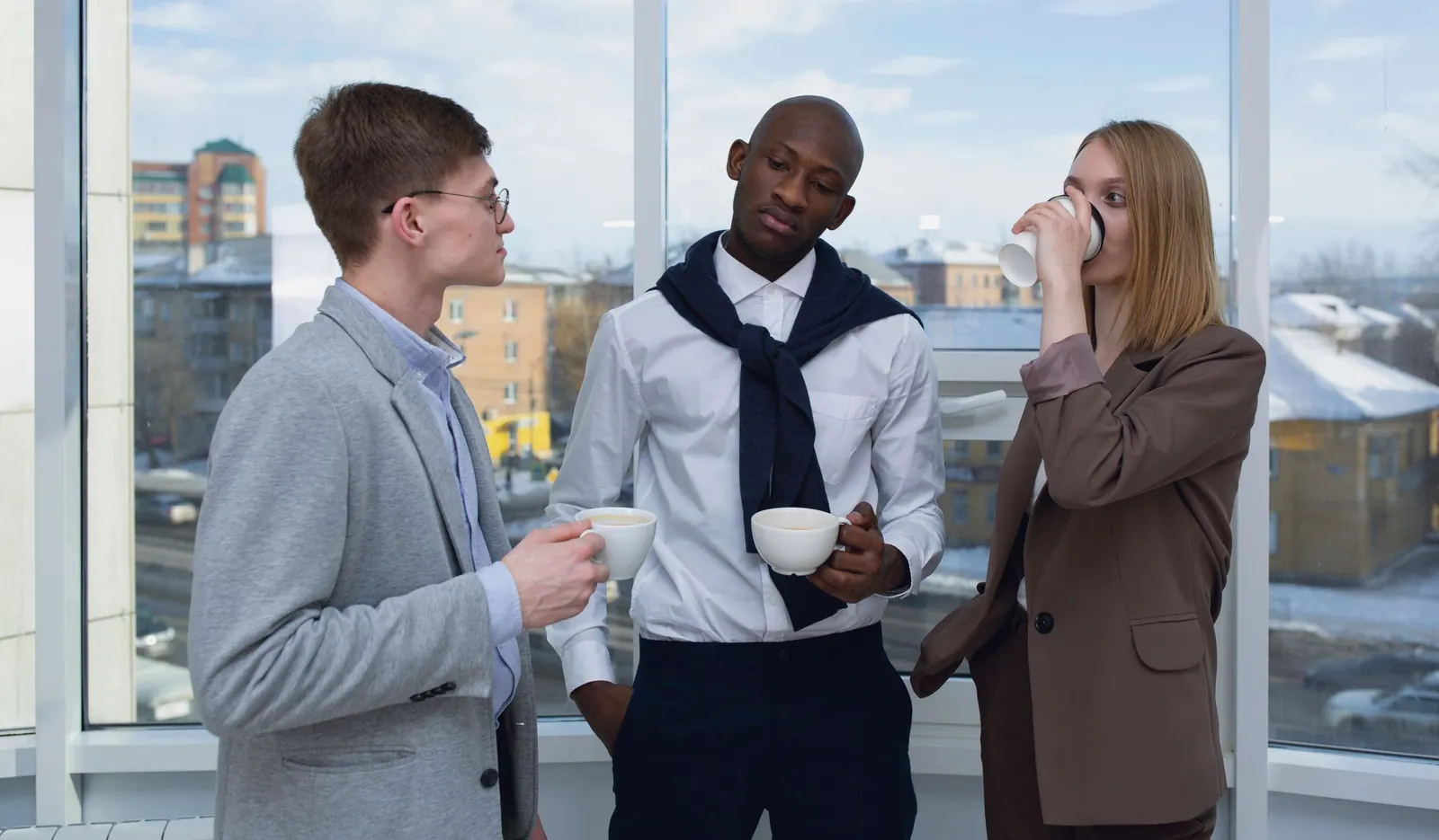 Three colleagues take a coffee break and talk by a bright window.