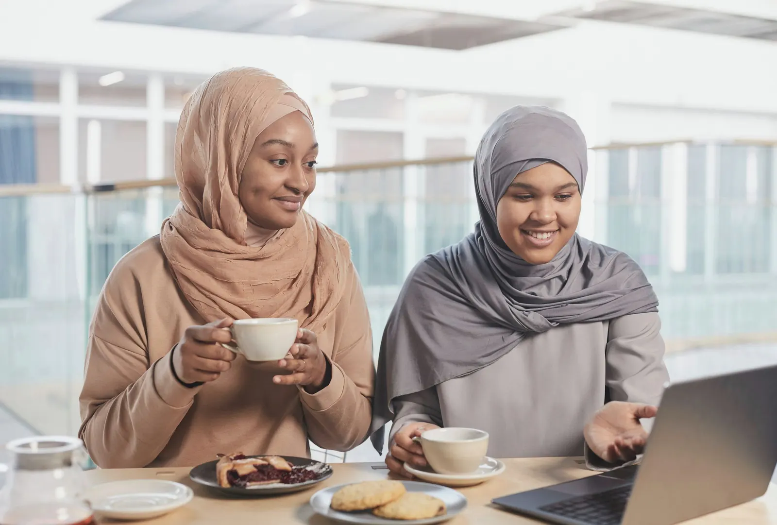 Two colleagues share coffee and pastries over a laptop.