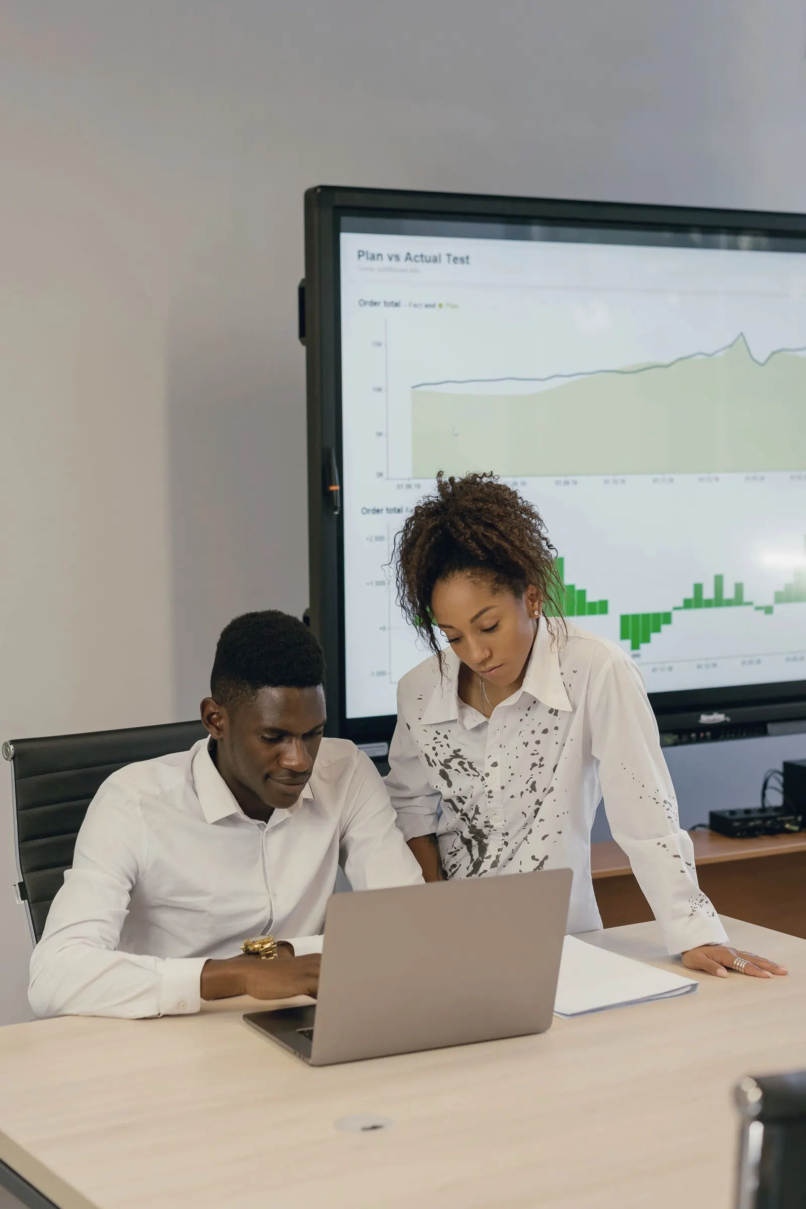 Two colleagues review a chart on a wall display and a laptop in a bright office.
