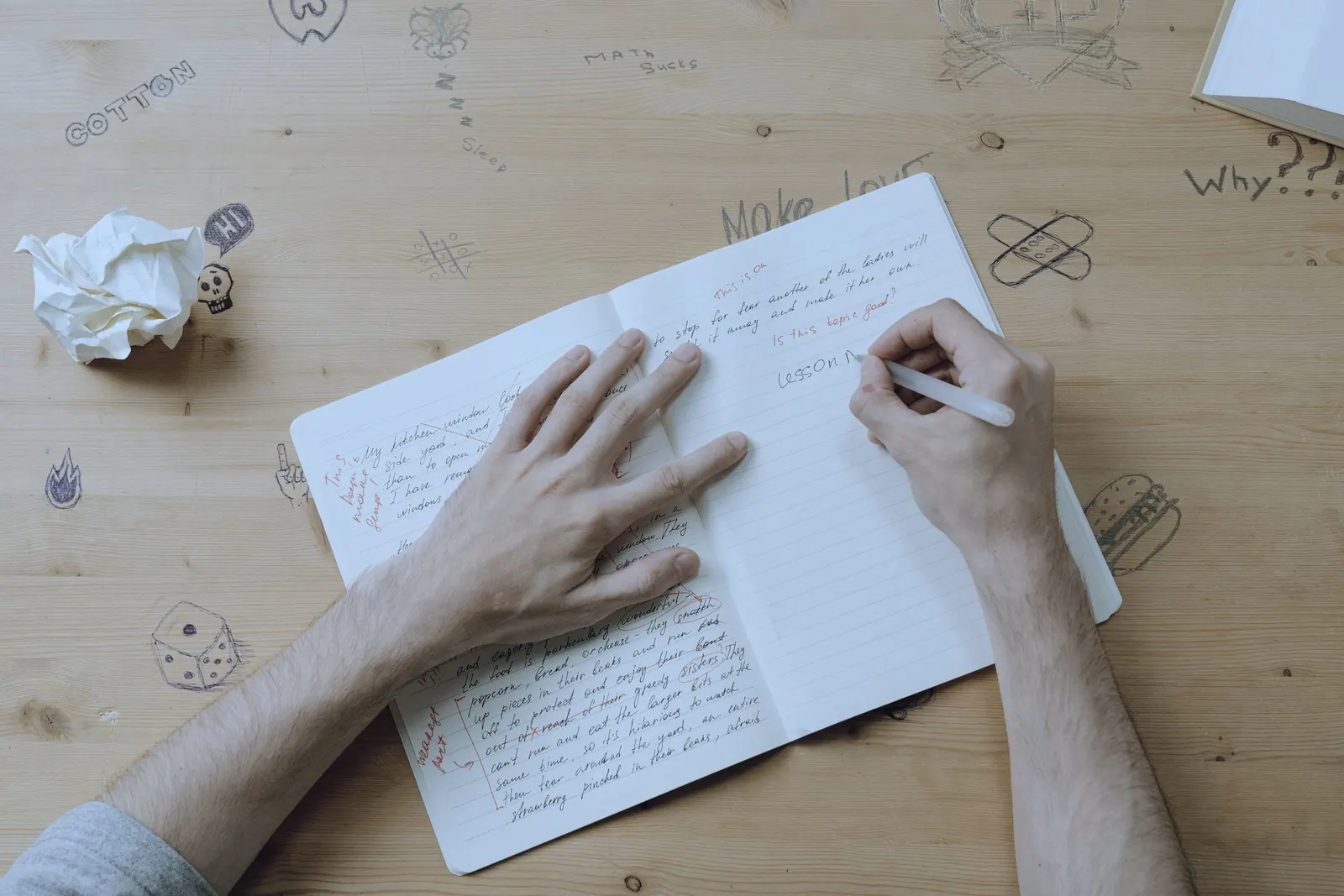Hands write in a notebook margined with doodles on a wooden desk.