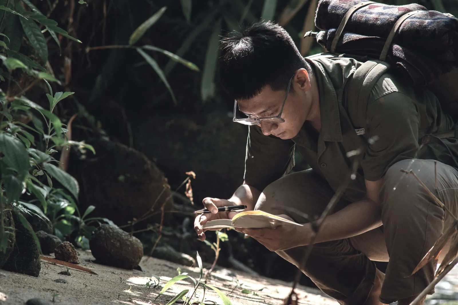A researcher crouches in the field, writing observations in a notebook.