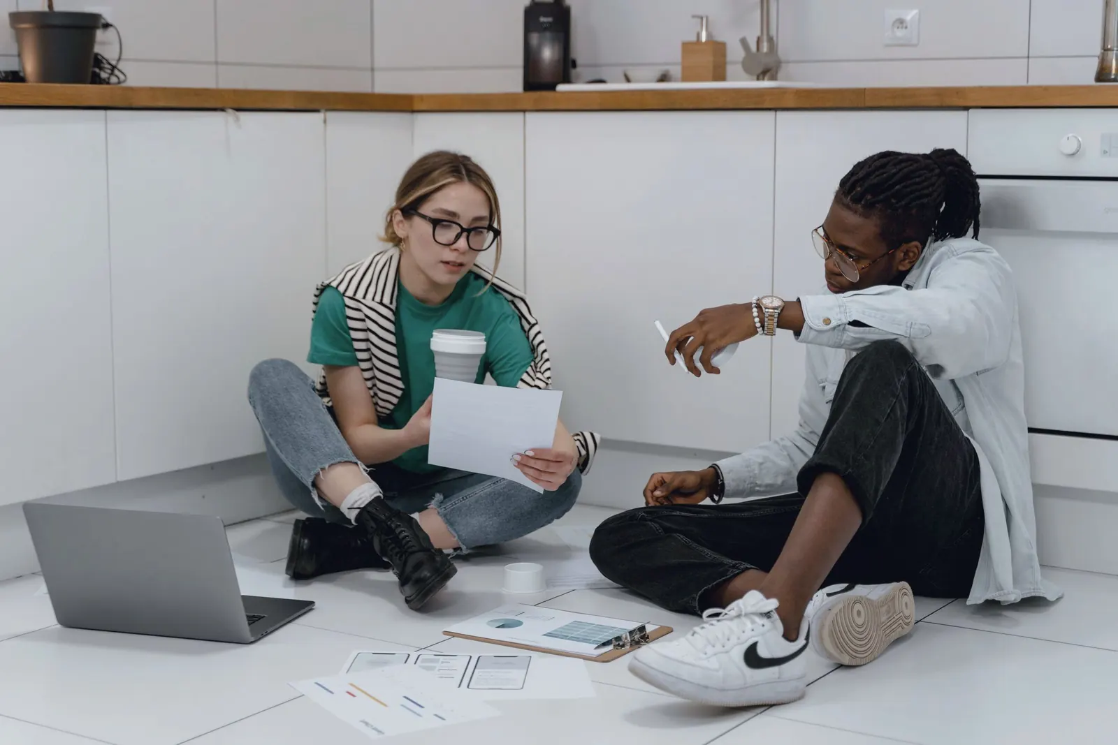 Two colleagues sit on a kitchen floor with papers and a laptop, planning.