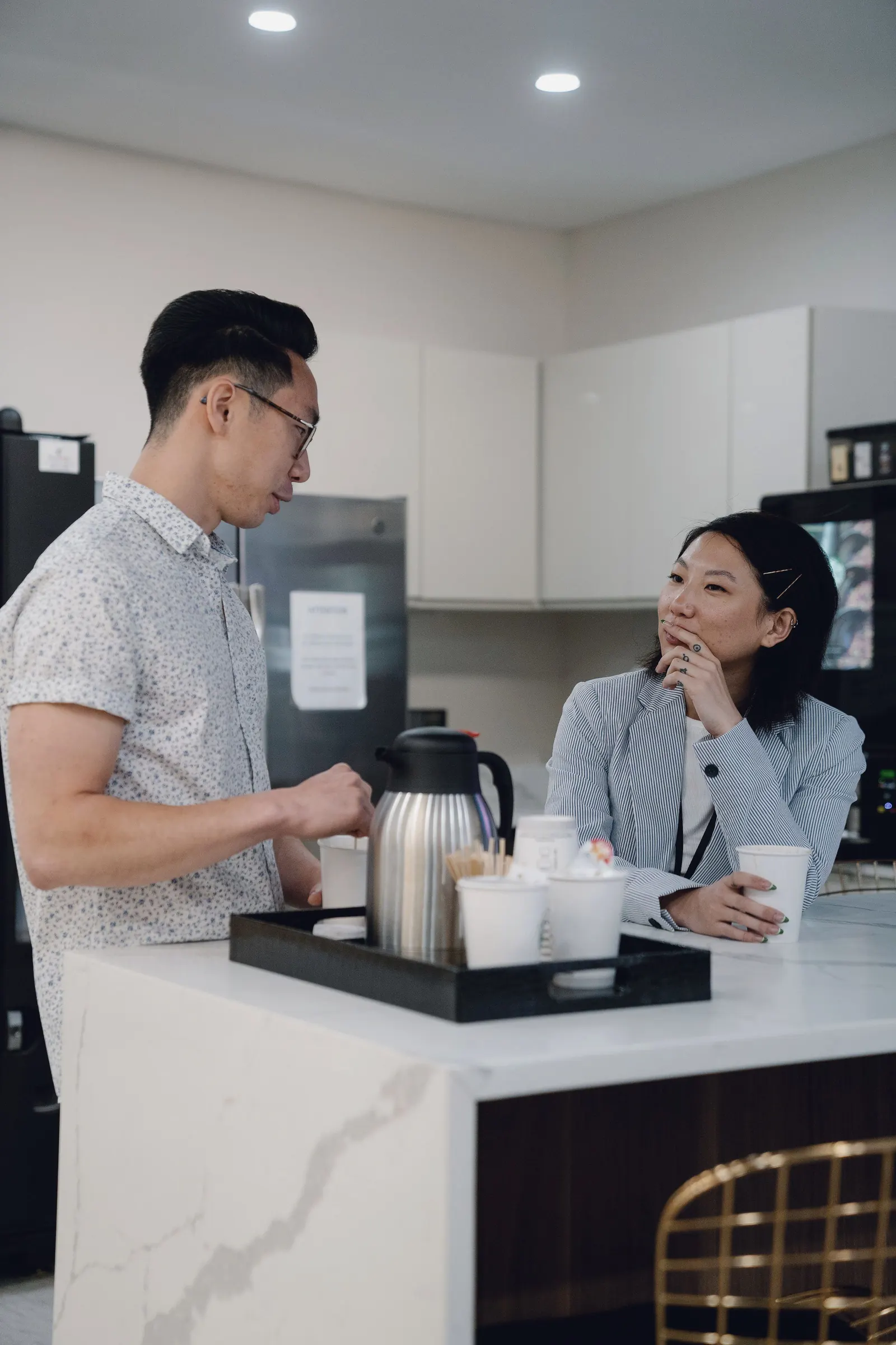 A colleague makes coffee while another smiles from the office kitchen table.