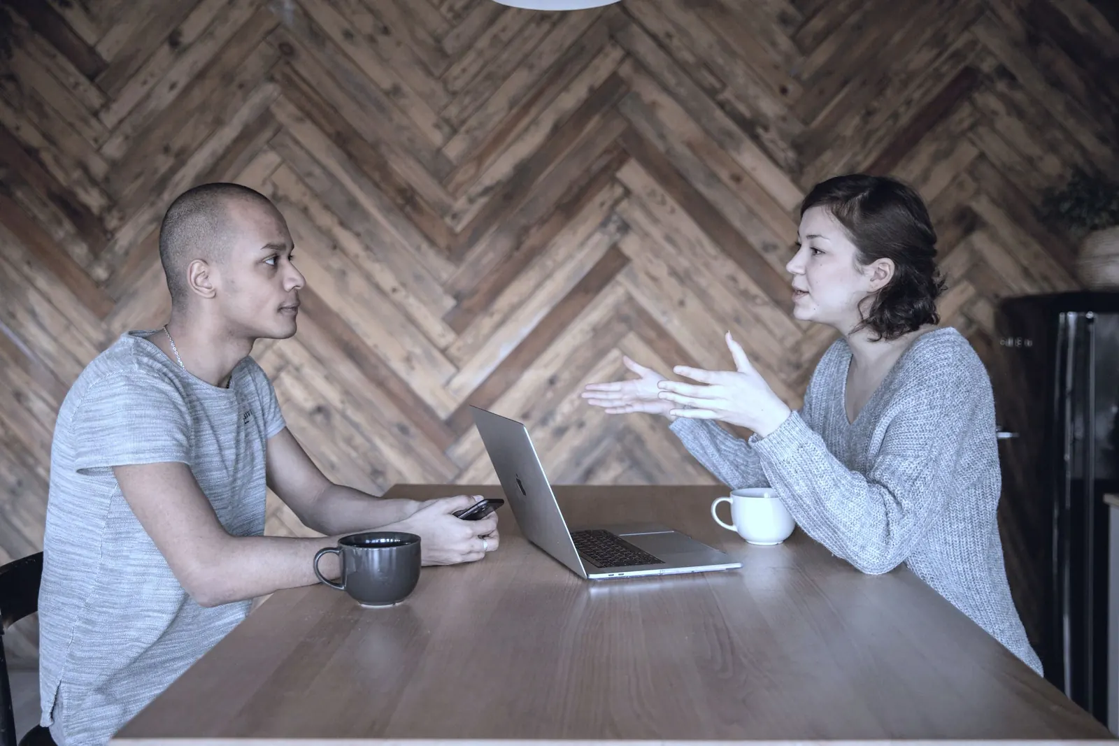 Two colleagues talk over mugs and a laptop at a wooden kitchen table.