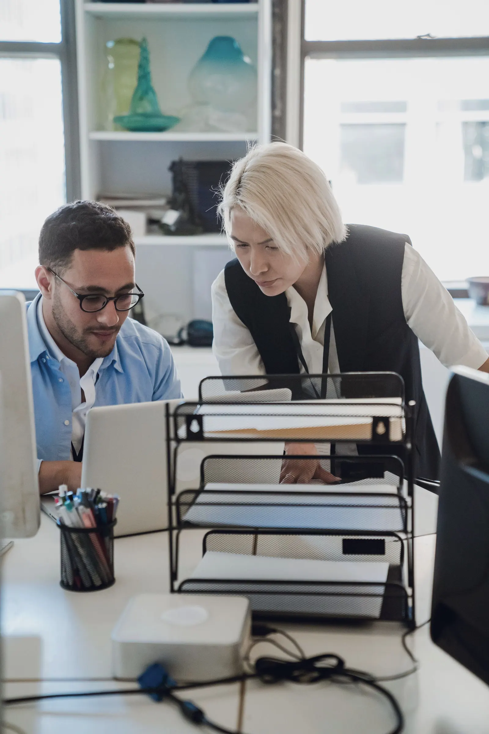 A colleague looks on as a coworker types at a laptop in a bright office.