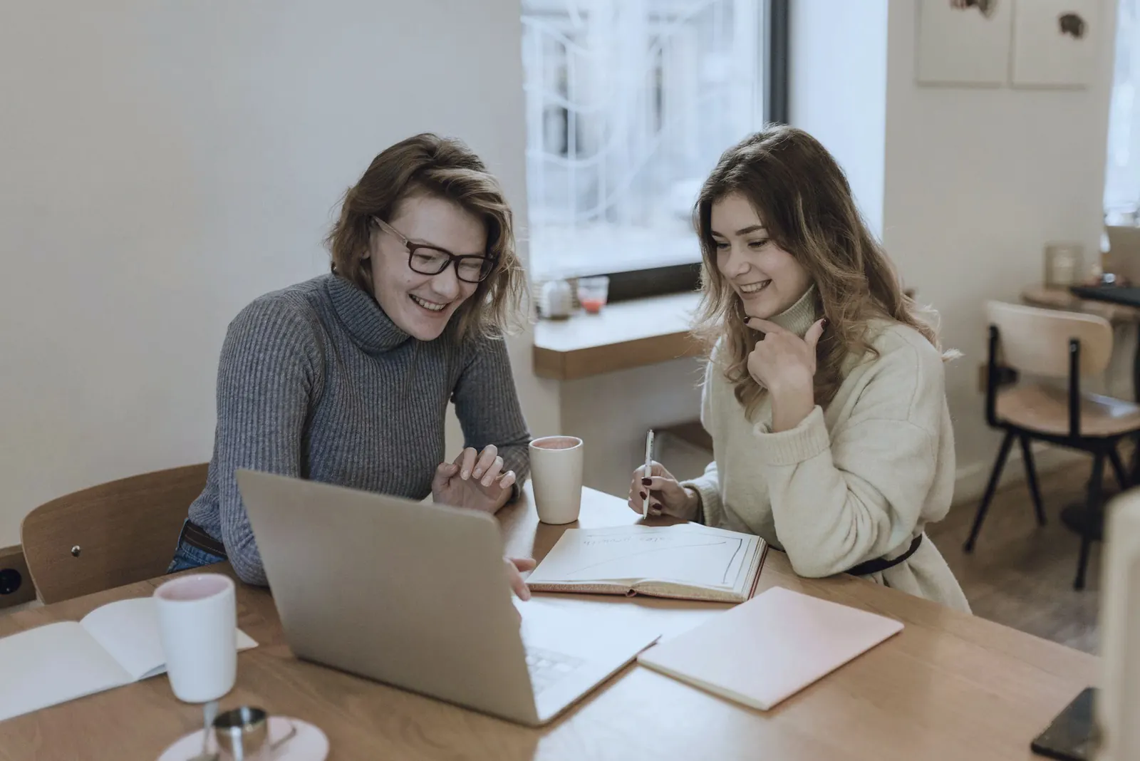 Two colleagues talk over a laptop, a notebook and a mug on a wooden table.