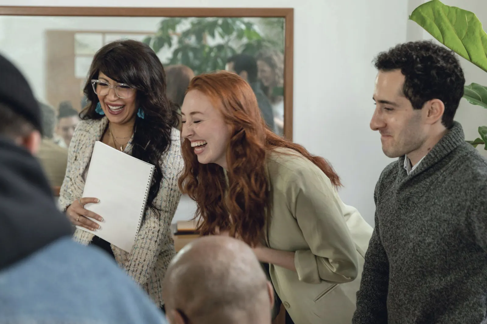 Colleagues laugh together over a notebook among the plants of a bright office.