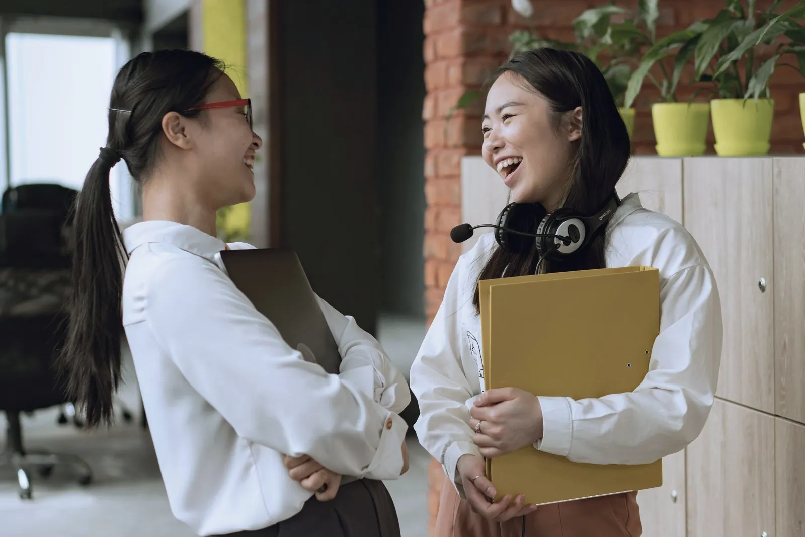 Two colleagues laugh together in a bright office, folders in their arms.
