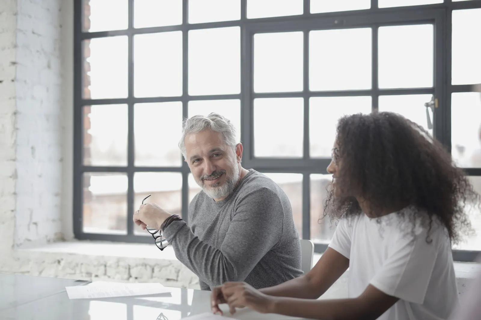 A senior colleague smiles as a younger coworker talks, beside a tall window.