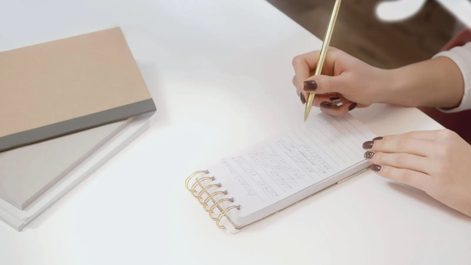 A hand writes in a spiral notebook on a white desk.