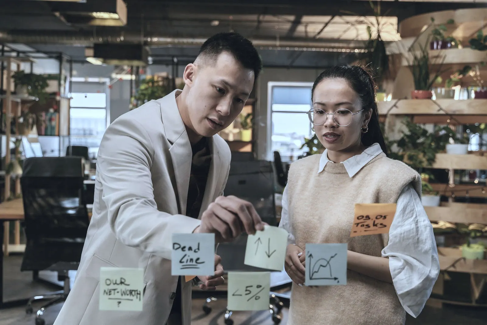 Two colleagues place sticky notes on a glass wall in a warm loft office.