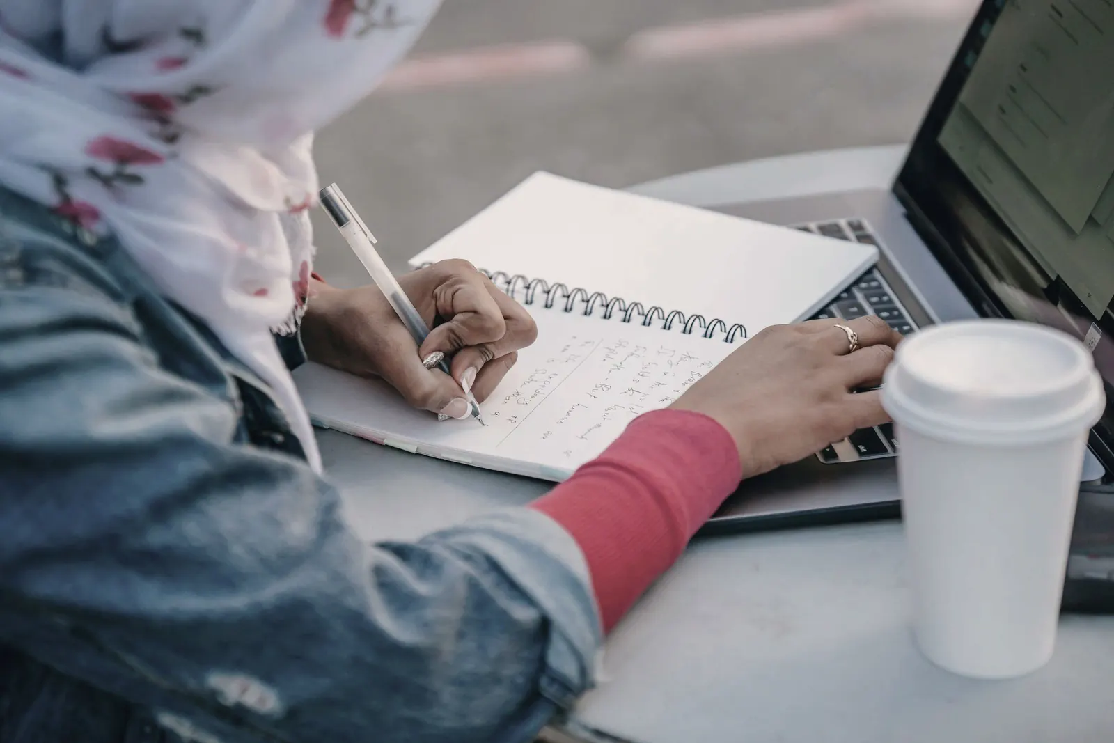 A hand writes in a notebook beside a laptop and a paper cup.
