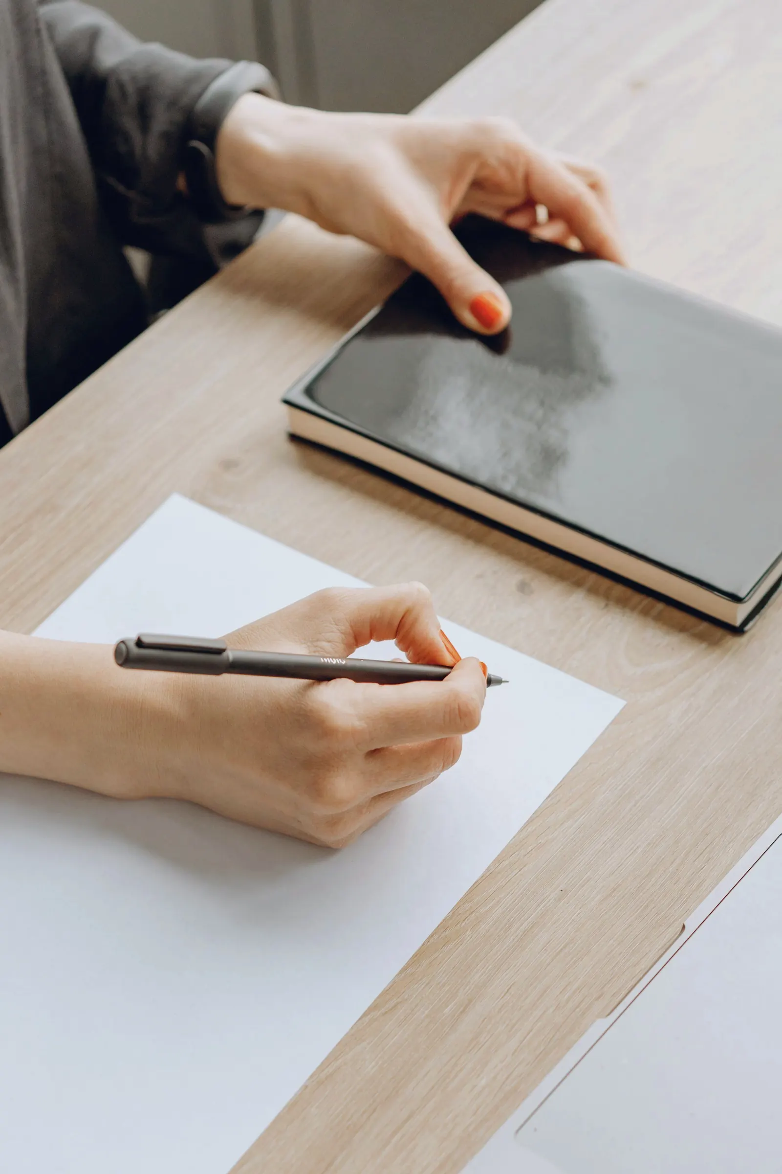 A hand writes on a sheet of paper beside a closed notebook.