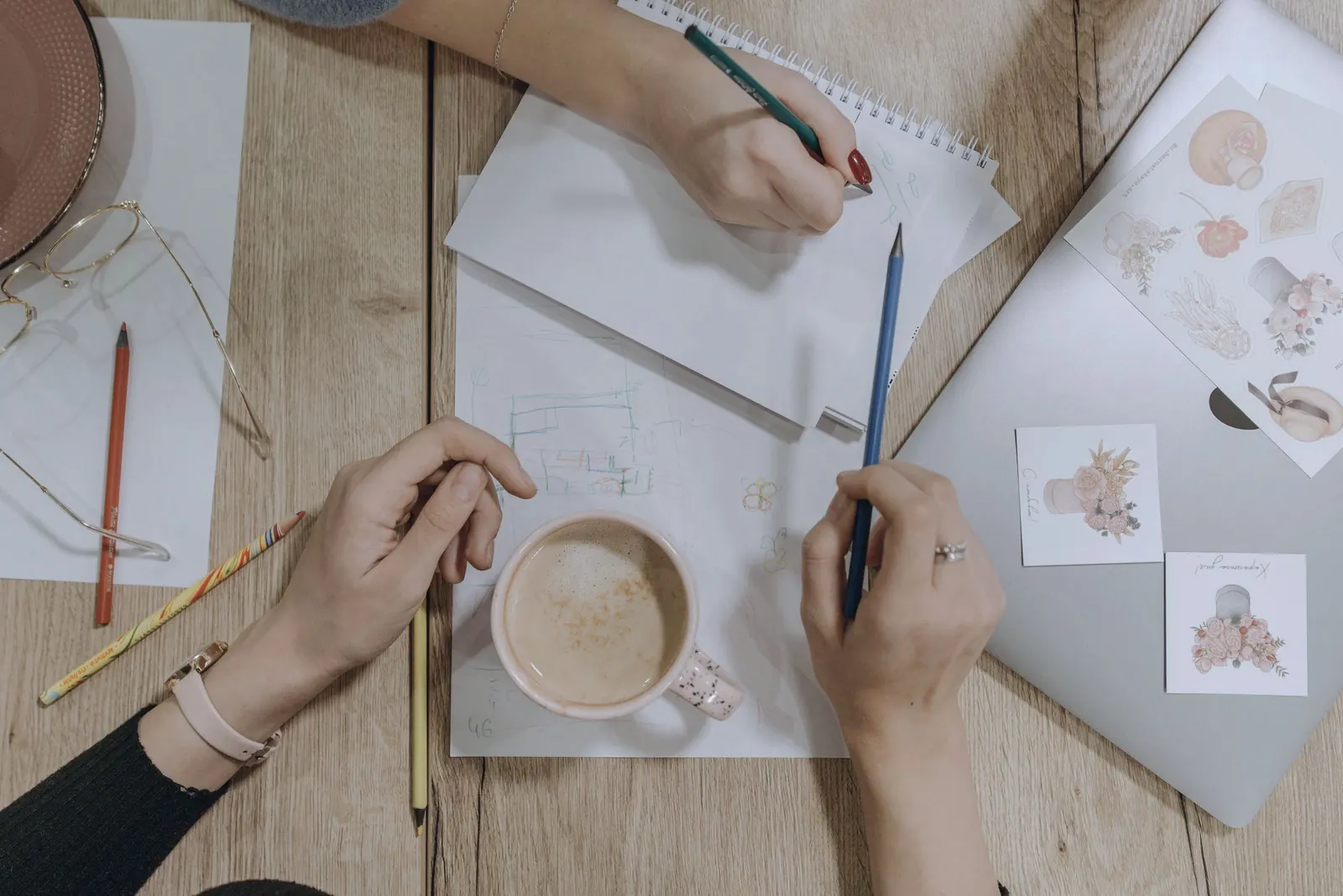 Two pairs of hands sketch on paper beside a cup of coffee, seen from above.