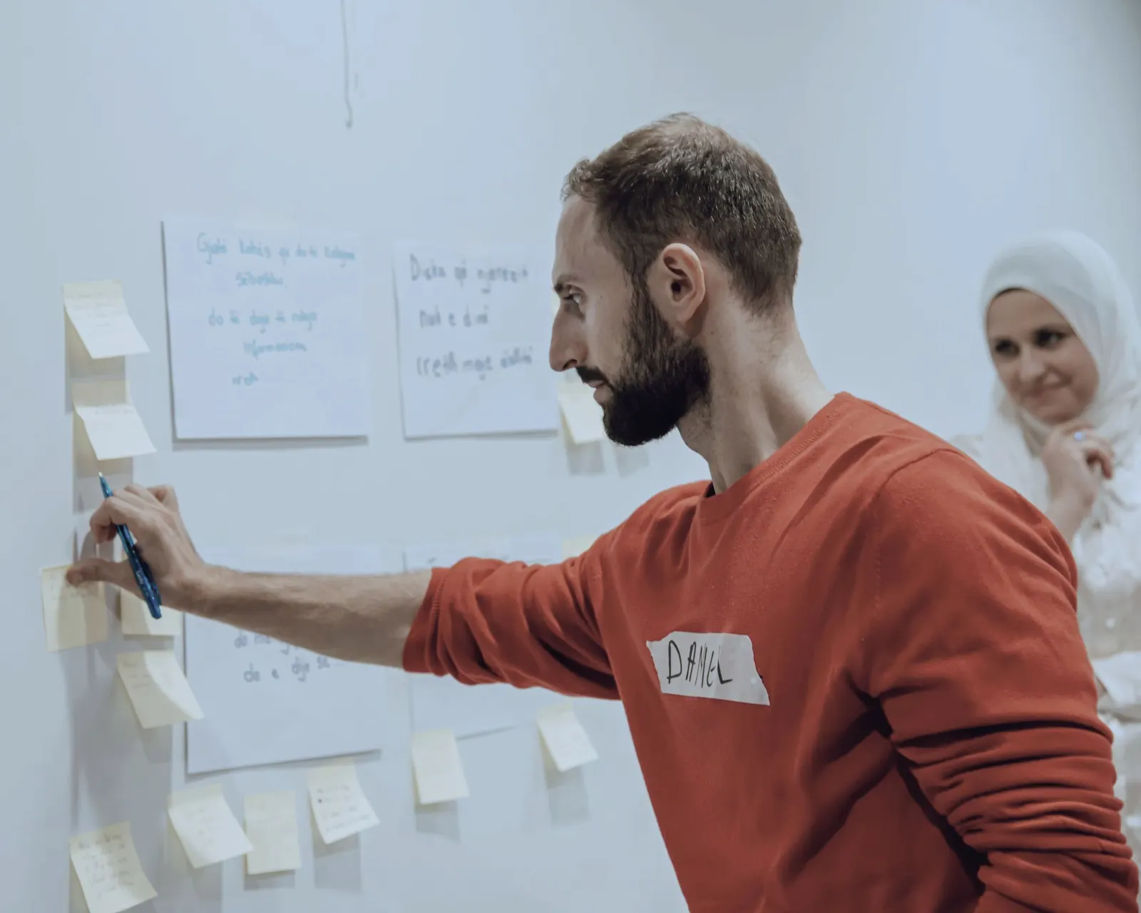 A colleague writes on notes stuck to a wall as a coworker looks on.