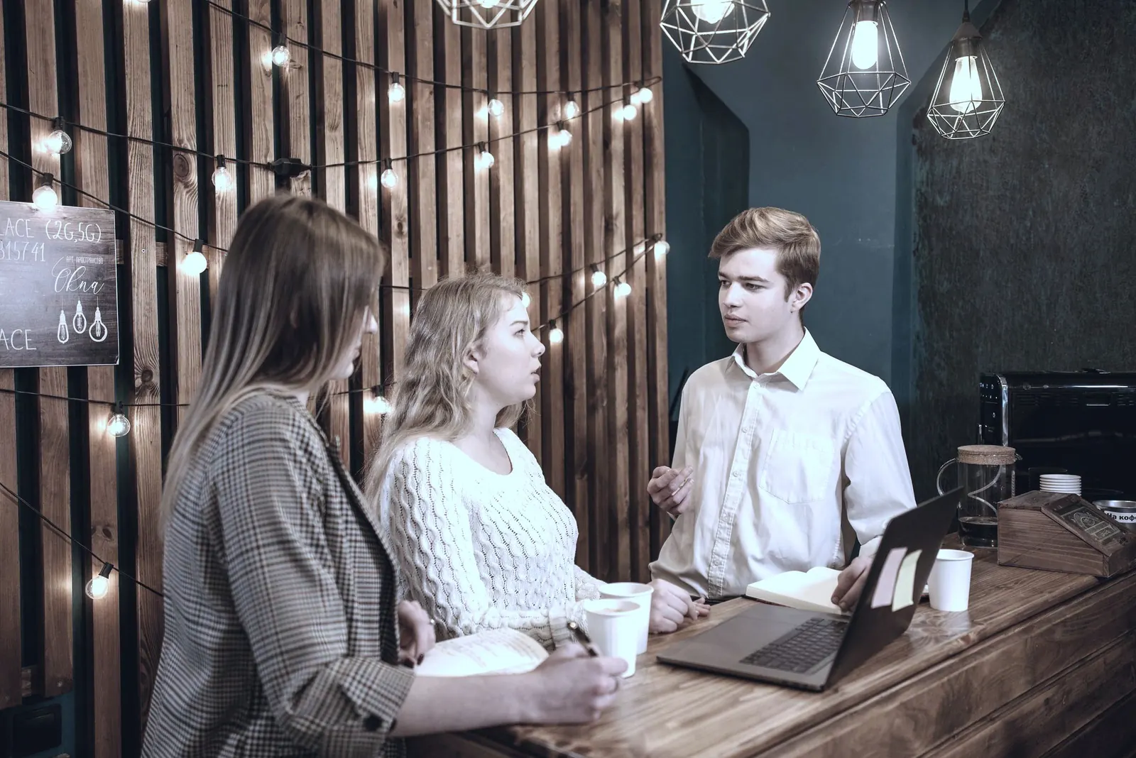 Three colleagues talk over a laptop and mugs at a wooden counter under string lights.