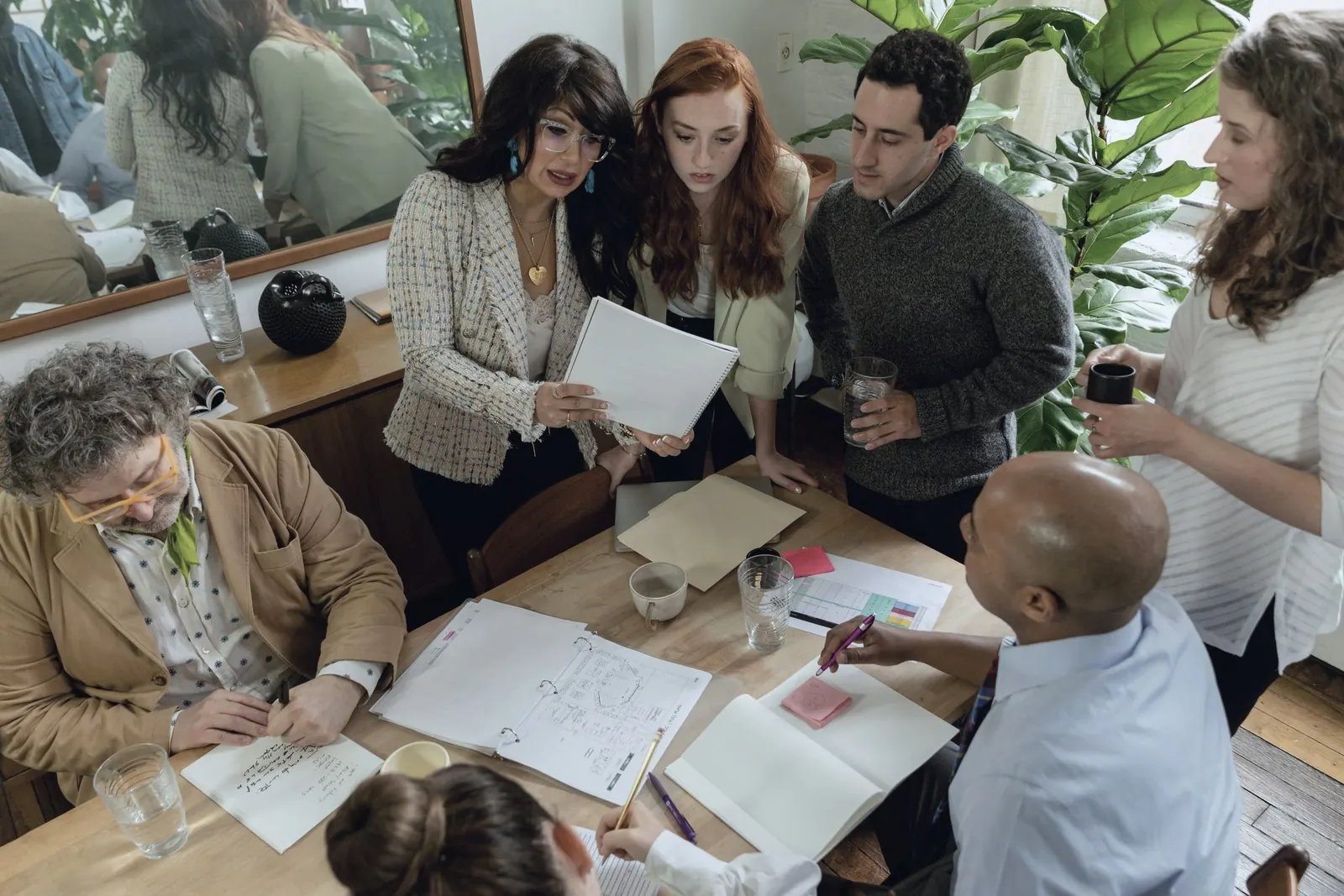 Colleagues sit around a wooden table with notebooks and sketches, seen from above.