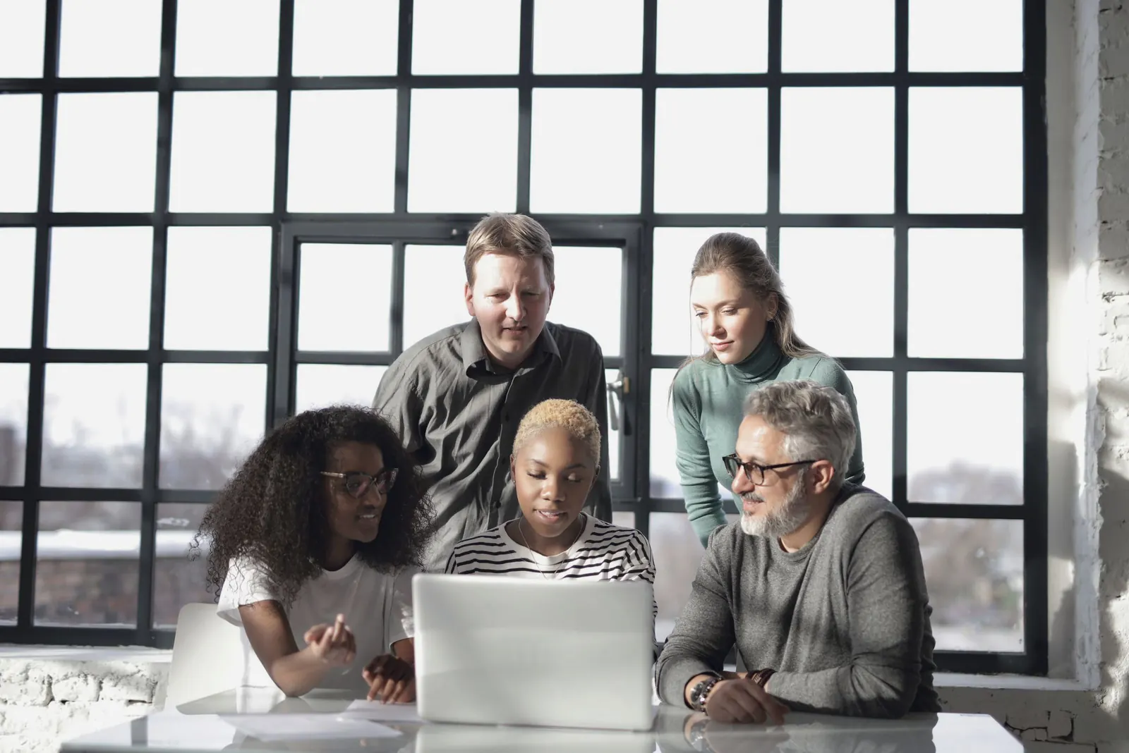 Five colleagues gather around a laptop in front of a tall gridded window.
