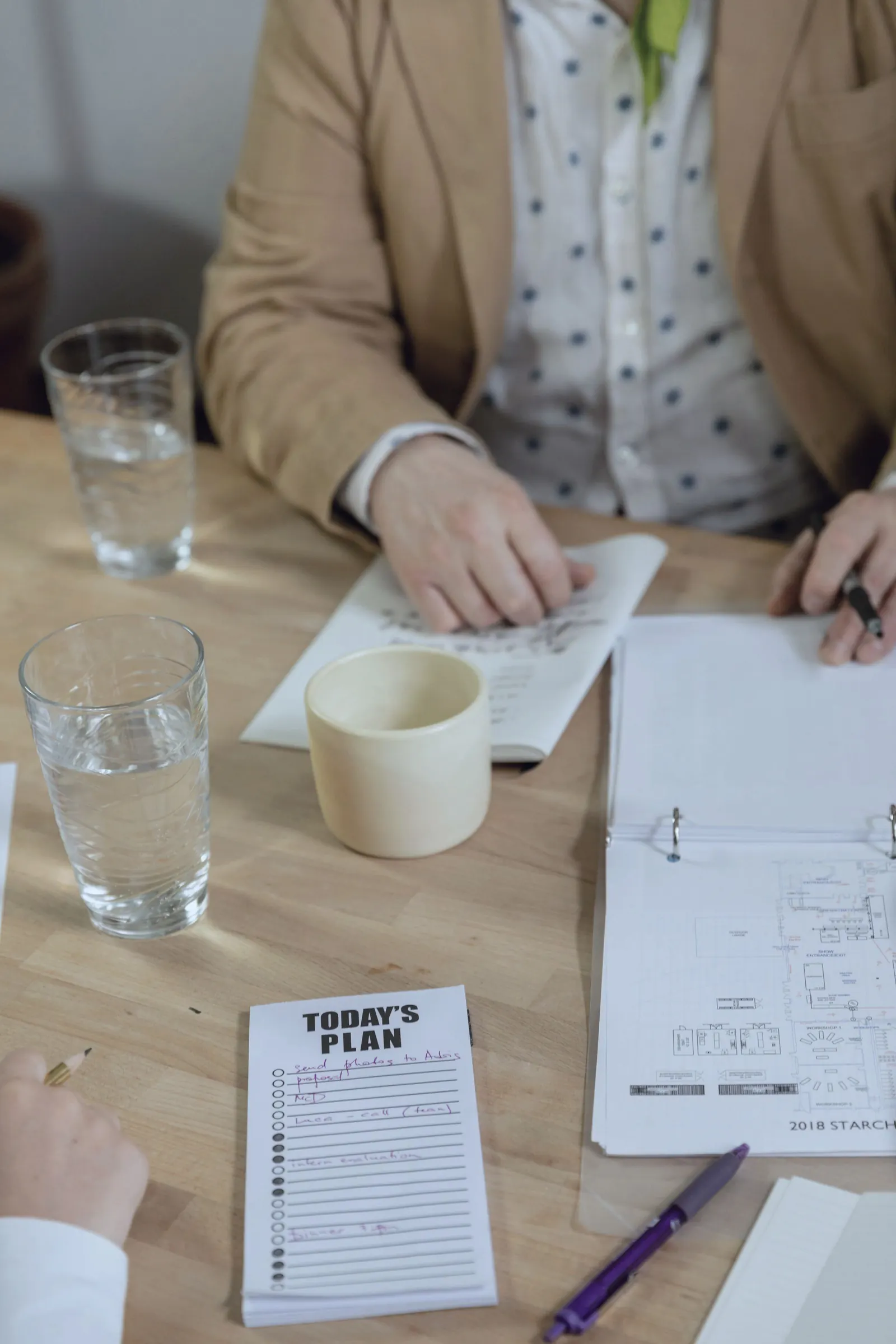 Hands fill in a daily plan and a notebook at a table with mugs and glasses of water.