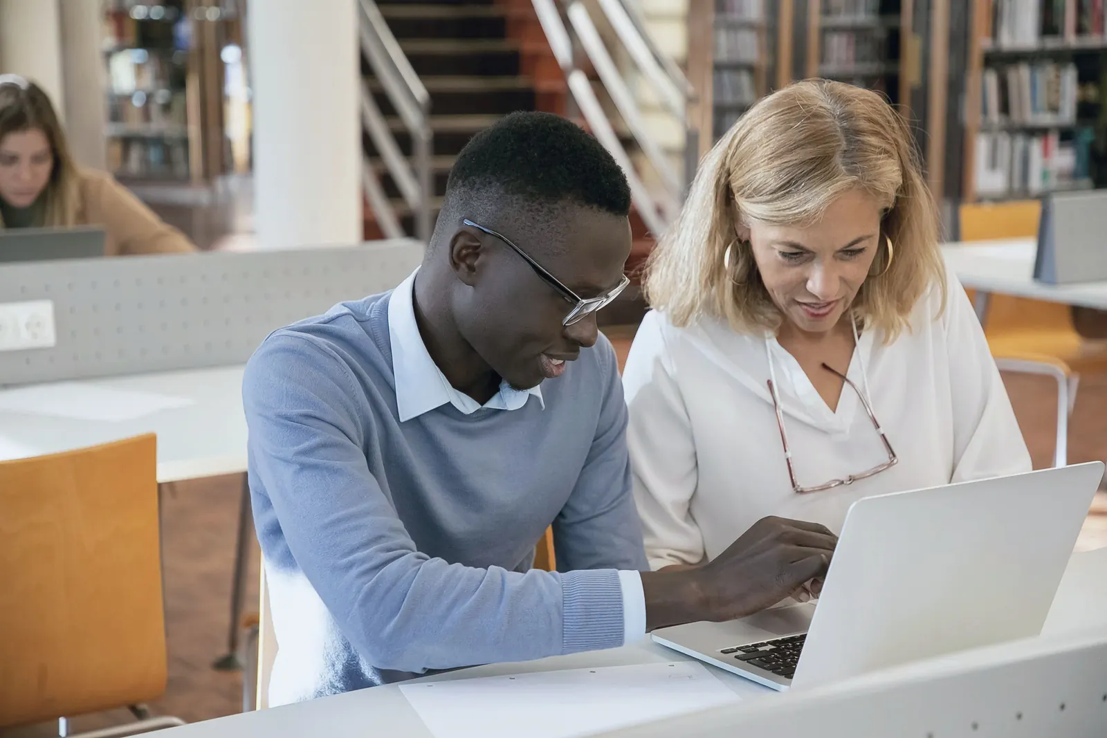A tutor and a student work through a problem at a laptop in a university library.