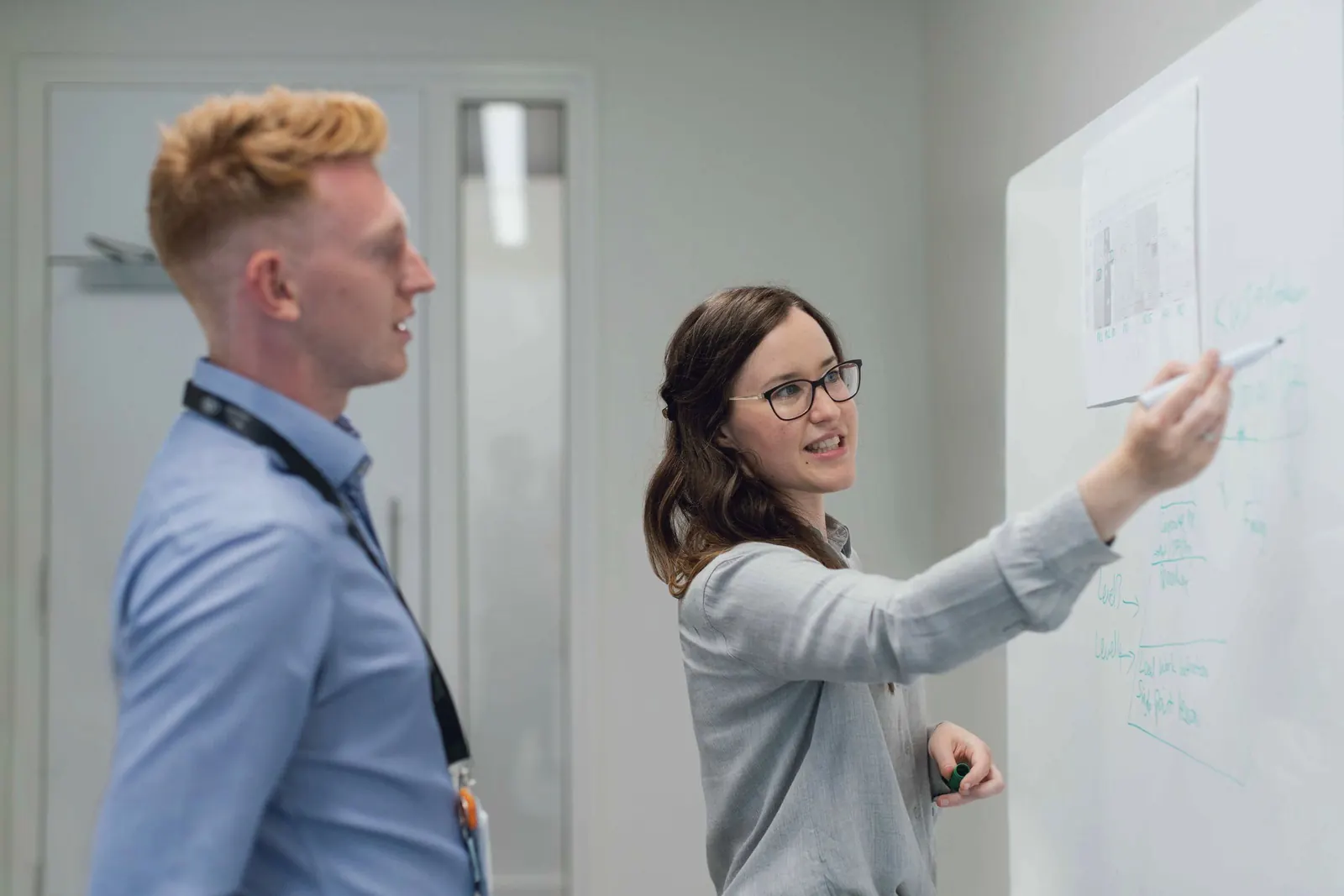 Two colleagues talk through a problem at a whiteboard.