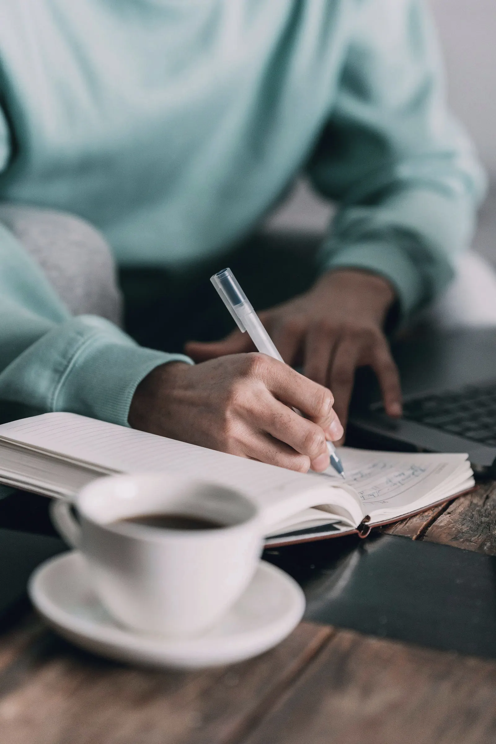 A colleague writes notes in a notebook beside a cup of coffee and a laptop.